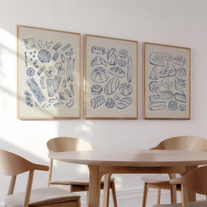 Puede incluir: Tres impresiones enmarcadas con ilustraciones de pasta, pan y queso cuelgan sobre una mesa y sillas de madera. Las impresiones presentan dibujos lineales azules detallados sobre un fondo crema, cada uno en un marco de madera clara.