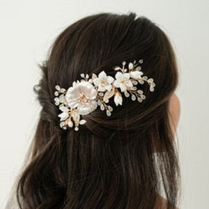 May include: A white and gold floral hair comb with pearl accents. The comb is worn in a woman's dark brown hair.