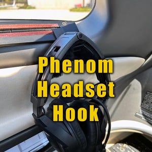 May include: A black headset hook for a Phenom aircraft. The hook is attached to the top of the aircraft's window frame. The headset is hanging from the hook.