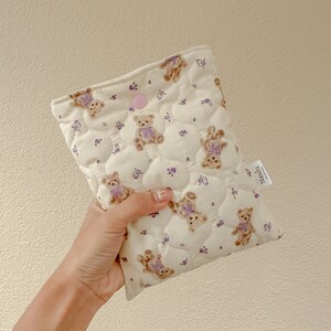 Op de afbeelding: Een gestikte cr&egrave;mekleurige tas met een roze knoopsluiting. De tas is versierd met een herhalend patroon van bruine teddyberen die paarse strikjes dragen en kleine paarse bloemen.