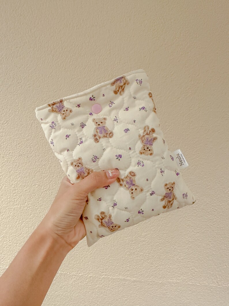 Op de afbeelding: Een gestikte cr&egrave;mekleurige tas met een roze knoopsluiting. De tas is versierd met een herhalend patroon van bruine teddyberen die paarse strikjes dragen en kleine paarse bloemen.