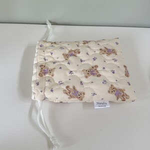 Op de afbeelding: Een klein, cr&egrave;mekleurig trekkoordtasje met een gewatteerde textuur. De tas is versierd met een herhalend patroon van bruine teddyberen die paarse strikjes en kleine paarse bloemetjes dragen. De tas heeft een wit trekkoord en een wit label met de tekst "Neulu Handmade".