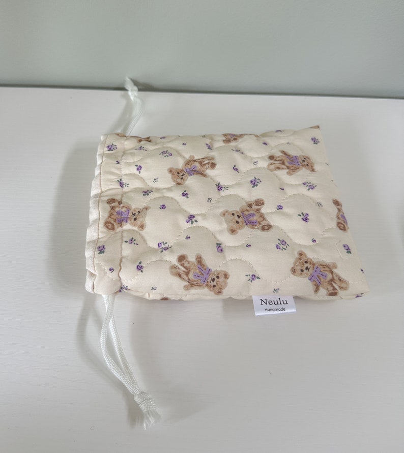 Op de afbeelding: Een klein, cr&egrave;mekleurig trekkoordtasje met een gewatteerde textuur. De tas is versierd met een herhalend patroon van bruine teddyberen die paarse strikjes en kleine paarse bloemetjes dragen. De tas heeft een wit trekkoord en een wit label met de tekst "Neulu Handmade".