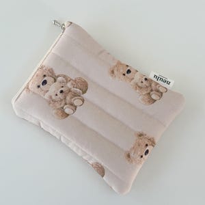 May include: A beige quilted zippered pouch with a brown teddy bear print. The pouch has a silver zipper pull and the word "neutr" printed on the side.
