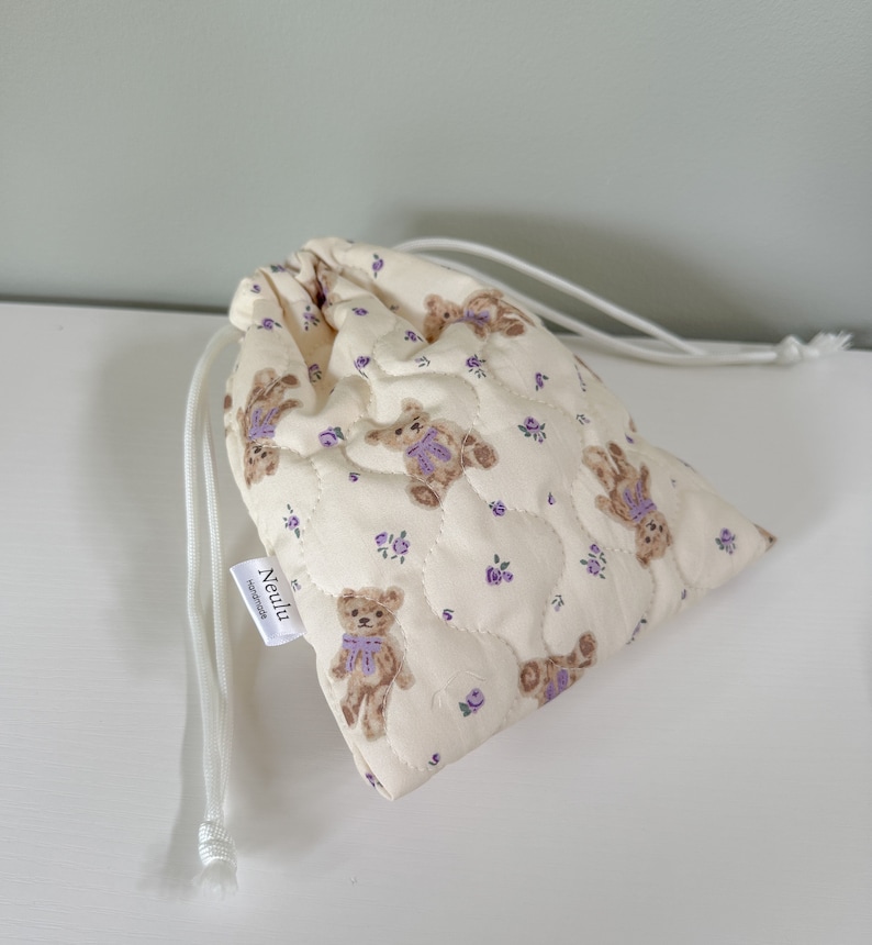 Op de afbeelding: Een witte trekkoordtas met een gewatteerde textuur en een patroon van bruine teddyberen met paarse strikken en kleine paarse bloemen. De tas heeft een wit trekkoord en een wit label met de tekst "Neulu" en het woord "handgemaakt" eronder.
