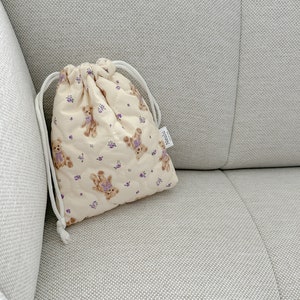 Op de afbeelding: Een klein, cr&egrave;mekleurig trekkoordtasje met een gewatteerde textuur. De tas heeft een herhalend patroon van bruine teddyberen met paarse bloemen. De tas heeft een wit trekkoord en een klein wit label met de tekst "Nunu" in zwarte letters.