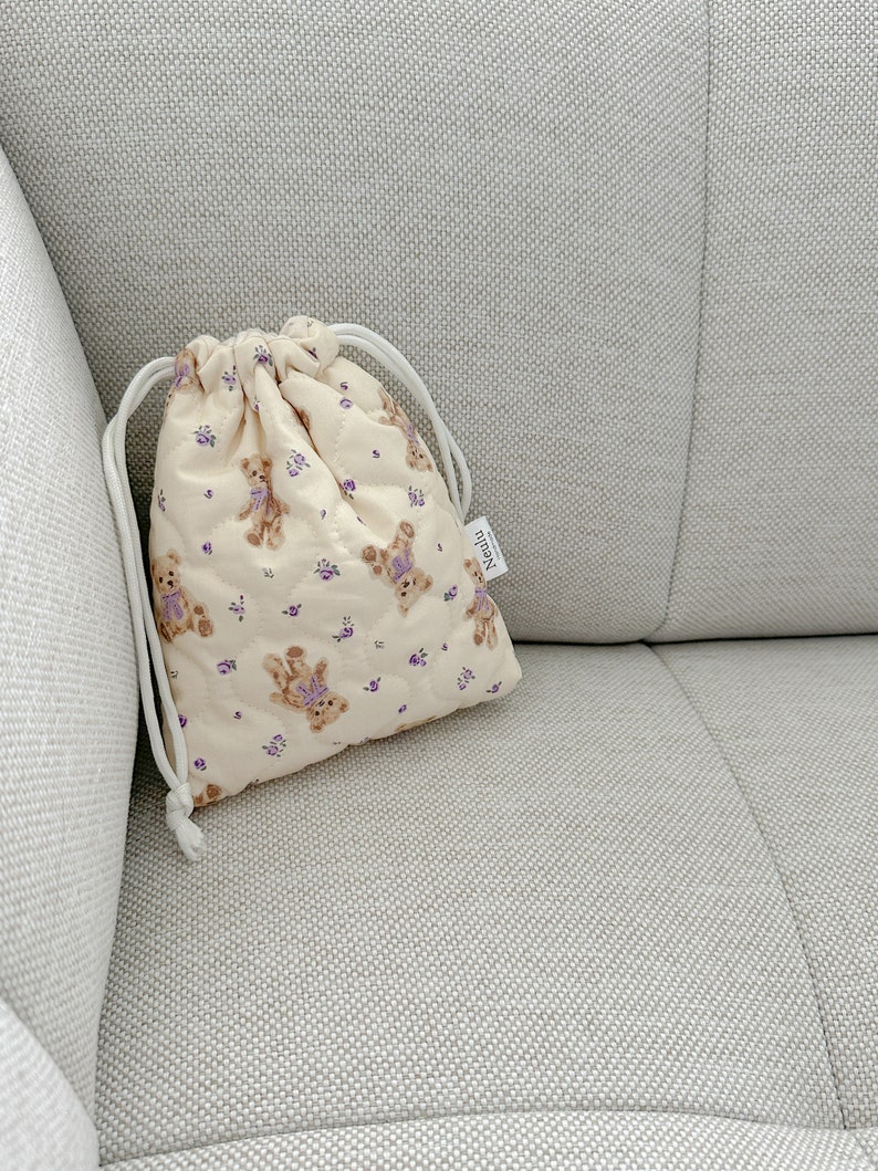 Op de afbeelding: Een klein, cr&egrave;mekleurig trekkoordtasje met een gewatteerde textuur. De tas heeft een herhalend patroon van bruine teddyberen met paarse bloemen. De tas heeft een wit trekkoord en een klein wit label met de tekst "Nunu" in zwarte letters.