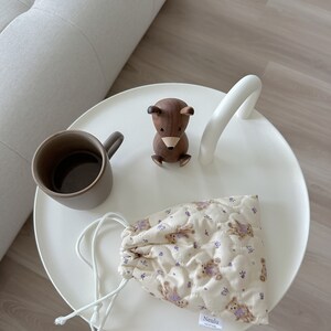 Op de afbeelding: Een witte tafel met een houten berenbeeldje, een bruine mok en een klein beige trekkoordtasje met een paars bloemenpatroon en de tekst "Neulu handmade" op de bodem.