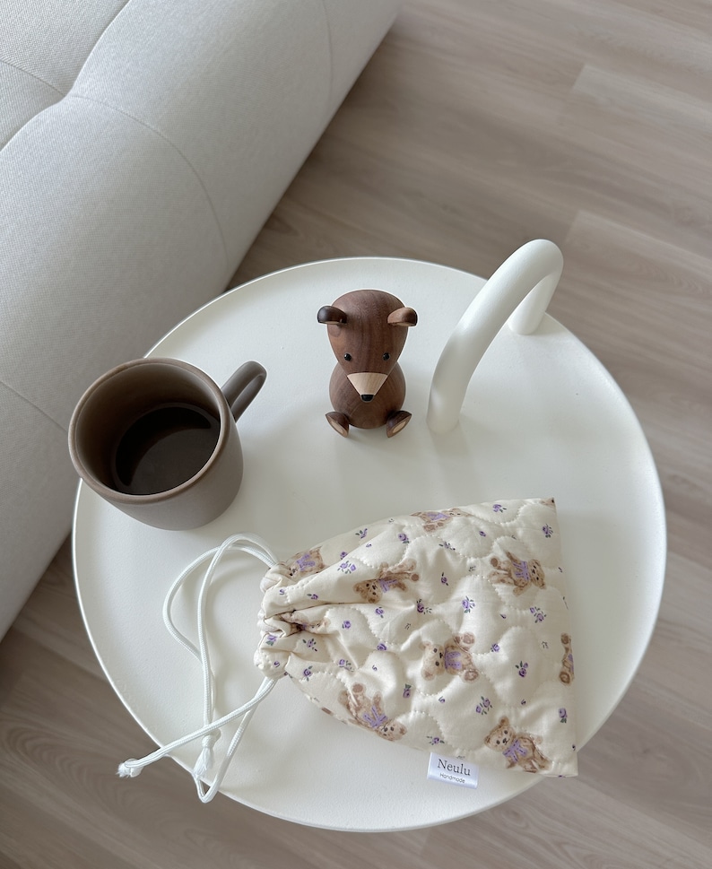 Op de afbeelding: Een witte tafel met een houten berenbeeldje, een bruine mok en een klein beige trekkoordtasje met een paars bloemenpatroon en de tekst "Neulu handmade" op de bodem.
