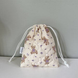 Op de afbeelding: Een klein, cr&egrave;mekleurig trekkoordtasje met een gewatteerde textuur. De tas is versierd met een herhalend patroon van bruine teddyberen en paarse bloemen. De tas heeft een wit trekkoord en een label met de tekst "Neulu".