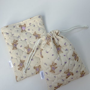 Op de afbeelding: Twee gevoerde stoffen tassen met een teddybeer- en bloemenprint. De tassen zijn beige met paarse bloemen en bruine teddyberen. De tassen hebben een trekkoord sluiting en een klein label met de tekst "Neulu".