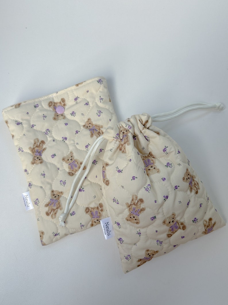 Op de afbeelding: Twee gevoerde stoffen tassen met een teddybeer- en bloemenprint. De tassen zijn beige met paarse bloemen en bruine teddyberen. De tassen hebben een trekkoord sluiting en een klein label met de tekst "Neulu".