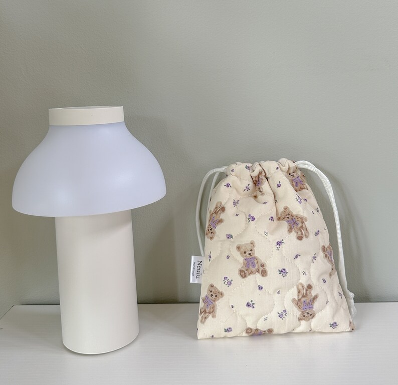 Op de afbeelding: Een witte, gewatteerde trekkoordtas met een beige achtergrond en een patroon van bruine teddyberen die paarse strikken dragen. De tas heeft een wit trekkoord en een wit label met het woord "Neulu" erop gedrukt.