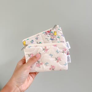 May include: Two quilted fabric zipper pouches. The top pouch has a floral pattern with small yellow, pink, and blue flowers. The bottom pouch has a white background with a pattern of small animals and flowers. Both pouches have a white label with the word "remarkable" printed in black.