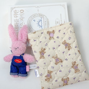 Op de afbeelding: Een kleine roze pluche teddybeer met een spijkerbroek overall ligt naast een beige gewatteerde tas met een bruine beerprint. De tas heeft een paarse knoopsluiting en een wit label met de tekst "Neulu".