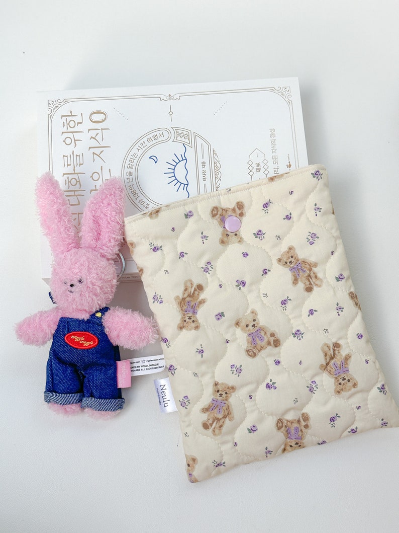 Op de afbeelding: Een kleine roze pluche teddybeer met een spijkerbroek overall ligt naast een beige gewatteerde tas met een bruine beerprint. De tas heeft een paarse knoopsluiting en een wit label met de tekst "Neulu".