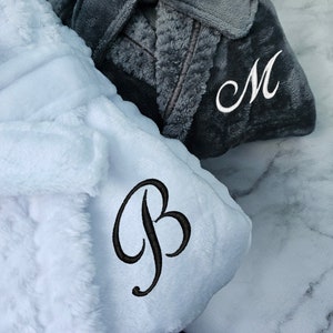 May include: Two plush robes, one gray and one white, with embroidered letters. The gray robe has an "M" embroidered on it, and the white robe has a "B" embroidered on it.