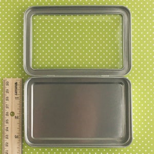 May include: An open, rectangular metal tin with a clear plastic lid. A ruler is placed next to the tin, showing measurements in inches. The tin is on a green background with white polka dots.
