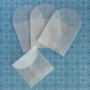 May include: Four translucent, off-white paper envelopes are arranged on a blue patterned surface. Three have an arched top, and one is square with a curved flap. The paper is thin and slightly textured.