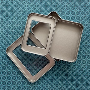 May include: A set of rectangular metal tins with rounded corners and a metallic finish. The set includes a base container, a lid with a clear rectangular window, and an additional frame piece, all displayed in an overlapping arrangement.