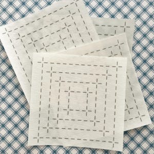 May include: Three white square fabric pieces with a grid pattern of dashed black lines. The fabric is layered on a blue and white plaid background. The stitching creates a square design.