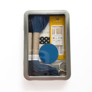 May include: A metal tin containing a sashiko stitching kit. The kit includes blue thread, needles, a thimble, and a blue fabric circle. The packaging features Japanese text and the brand name "Hidimari Sashiko."