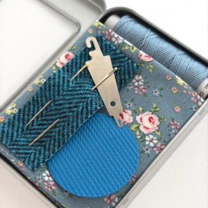 May include: A metal sewing kit tin containing a spool of light blue thread, a piece of blue floral fabric, a strip of teal and black herringbone fabric, a circular blue textured patch, and a silver metal needle threader with two needles.
