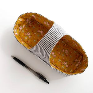 May include: An open fabric storage basket with a black and white pinstriped exterior and a mustard yellow interior lining featuring a small pink and green floral pattern. A black ballpoint pen is positioned next to the basket for scale.