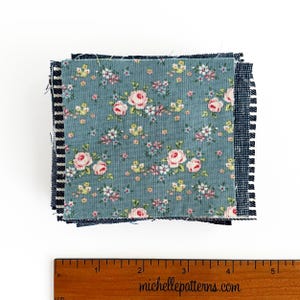 May include: A stack of fabric squares with floral and striped patterns. The top layer features a blue background with pink roses and small white and yellow flowers. The edges are frayed. A wooden ruler with inch measurements is at the bottom.