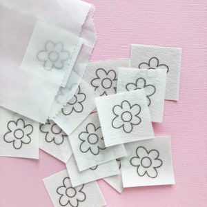 May include: A white paper bag with a pile of white paper squares with black outlined flower designs on a pink surface.