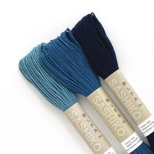 May include: Three skeins of Sashiko thread in shades of blue, from light to dark. Each skein is wrapped with a paper label that includes Japanese text and the word "Sashiko". The thread is 100% cotton.