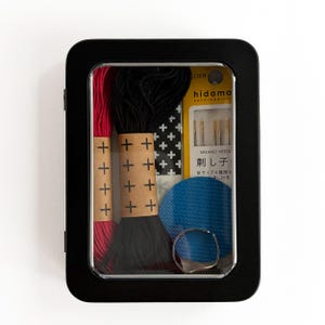 May include: A black rectangular metal tin with a clear window containing a Sashiko embroidery kit. Inside are red and black thread skeins with cross-patterned paper bands, a blue circular fabric patch, a silver thimble, and a pack of Sashiko needles.