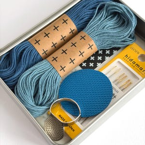 May include: A metal tin containing two skeins of blue embroidery thread wrapped in paper with a plus sign pattern, a blue circular textured disc, a silver metal thimble ring, and a pack of Lecien Hidamari Sashiko needles with yellow packaging.