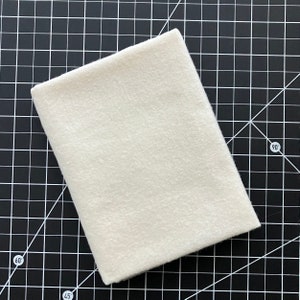 May include: A folded, off-white fabric square, possibly felt or a similar material, is positioned diagonally on a black cutting mat with white grid lines and angle markings. The fabric's texture appears soft and slightly fuzzy.