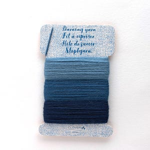 May include: A rectangular card wrapped with three shades of blue darning yarn, ranging from light to dark. The card features the text Darning yarn, Fil à repriser, Hilo de zurcir, Stoptgarn, and includes a single blue needle inserted at the top left.