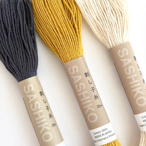 May include: Three spools of Sashiko thread in dark gray, mustard yellow, and off-white. Each spool is wrapped with a brown paper label featuring Japanese text and the brand name. The thread is 100% cotton and made in Japan.