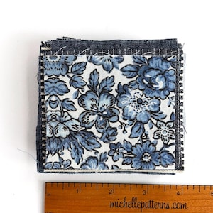 May include: A stack of square fabric swatches. The top swatch features a floral pattern in shades of blue and black on a white background. The edges are finished with a dark blue fabric. A wooden ruler with inch measurements is at the bottom.