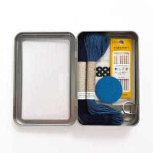 May include: An open metal tin containing a Sashiko embroidery kit. The kit includes two spools of blue thread, needles, a thimble, and a blue fabric circle. The kit also includes a small package of needles and a piece of fabric with a black and white cross pattern.