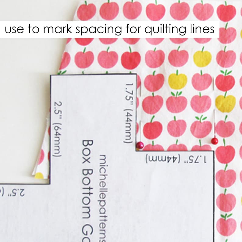 May include: A white paper template with markings for quilting lines. The template is labeled with measurements in inches and millimeters. The text "michellepatterns Box Bottom Go" is printed on the template. The template is placed on top of a fabric with a pink apple pattern.