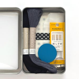 May include: Open metal tin containing a sashiko embroidery kit. The kit includes two spools of Sashiko thread in black and gray, needles, a blue fabric circle, and a white fabric with a cross pattern. The kit is made in Japan.