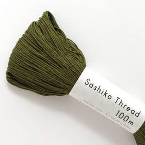 May include: A skein of olive green Sashiko thread made of 100% cotton. The white paper label features the text Sashiko Thread 100m, Cotton 100%, and Made in Japan, along with a faint geometric pattern printed on the surface of the paper label.