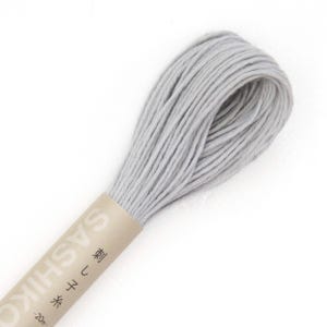 May include: A skein of light gray Sashiko thread, tightly wound and ready for use. The thread is wrapped with a tan paper label that has Japanese text and the word "SASHIKO" printed on it. The thread is approximately 20 meters in length.