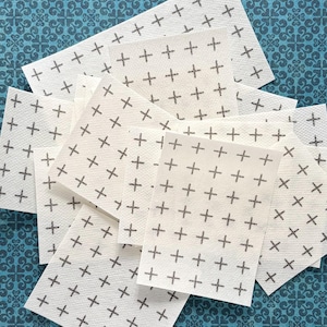 May include: A collection of rectangular off-white paper cards, each featuring a repeating pattern of small, dark gray plus signs arranged in a grid. The cards are layered and overlapping, displaying a consistent textured surface and uniform print.