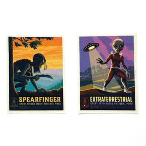 May include: Two vintage-style stickers. One features a creature labeled "SPEARFINGER" in front of a tree, with a sunset background. The other shows an alien and a UFO, labeled "EXTRATERRESTRIAL". Both include park names.