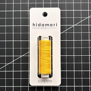 May include: A spool of bright yellow Sashiko thread by Hidamari, secured on a white rectangular cardboard display card. The card features the text 'hidamari SASHIKO' at the top and 'cosmo LECIEN CORPORATION' at the bottom, with a circular hole for hanging.