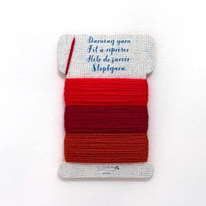May include: A rectangular card wrapped with three shades of red darning yarn. The card features the text Darning yarn, Fil à repriser, Hilo de zurcir, Stopfgarn, and Clover Japan. A single red needle is inserted into the top left of the card.
