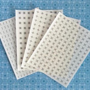 May include: Four rectangular sheets of off-white paper arranged in a fan shape. Each sheet features a distinct repeating pattern in dark gray: vertical lines, asterisks, plus signs, and multiplication signs, all organized in neat, uniform rows.