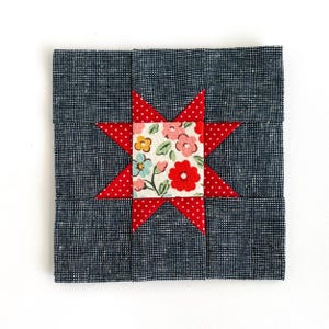 May include: A square fabric block with a central eight-pointed star design. The star is red with white polka dots, and the center features a floral pattern with pink, red, and blue flowers. The background is a dark blue fabric.