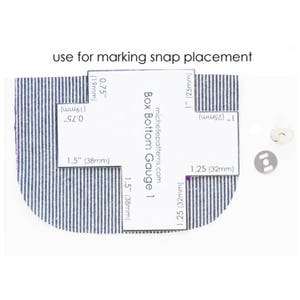 May include: A white guide with measurements in inches and millimeters for marking snap placement on fabric. The guide is labeled "Box Bottom Gauge 1" and includes the website address "michellepatterns.com".