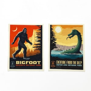 May include: Two rectangular Bigfoot and Creature From The Deep art prints. The Bigfoot print features a black silhouette against an orange sky. The Creature From The Deep print shows a green creature in a lake. Both prints have text.